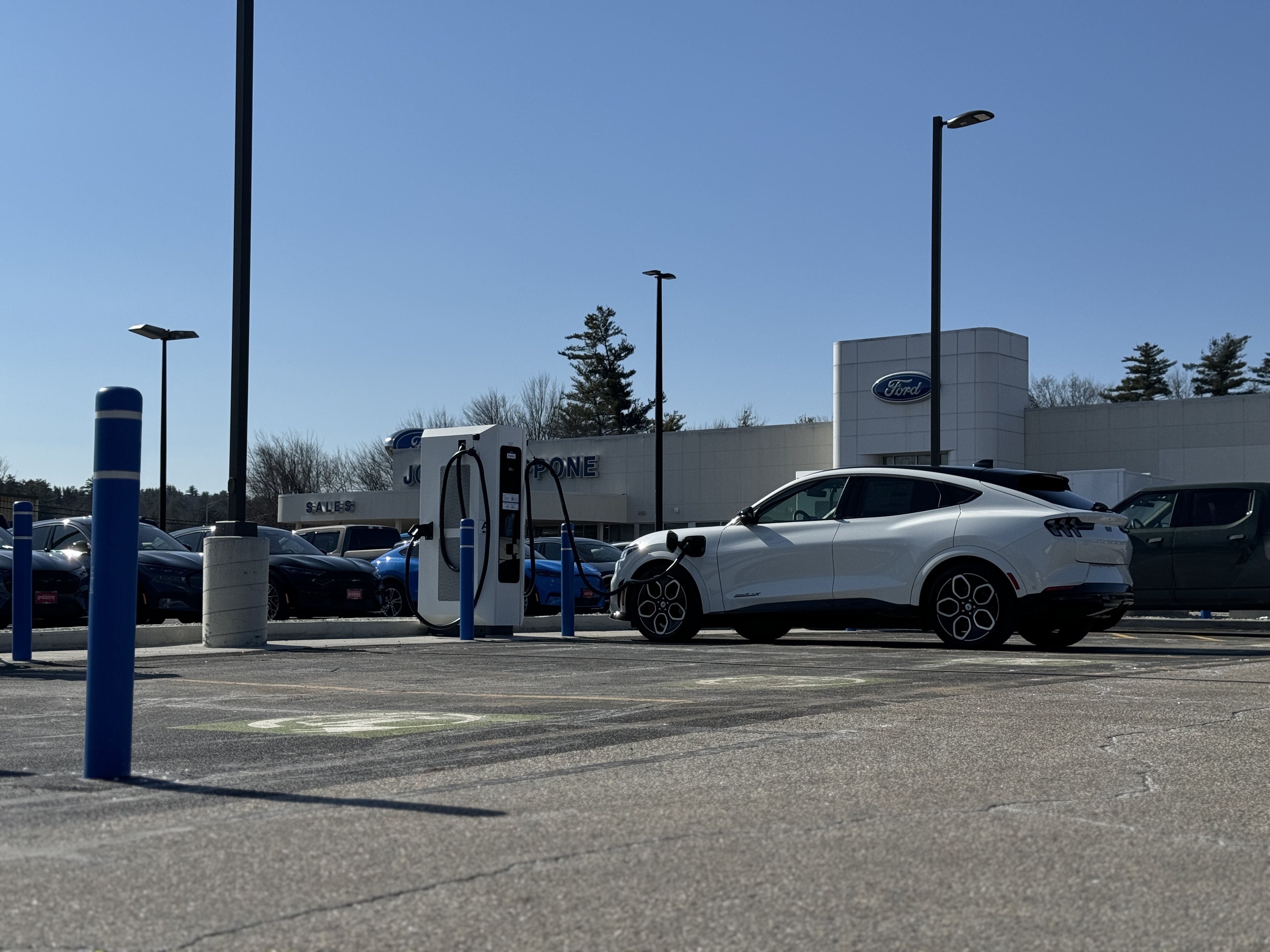 A white Ford Mustang Mach-E charges on a new charger in a Ford dealership parking lot.