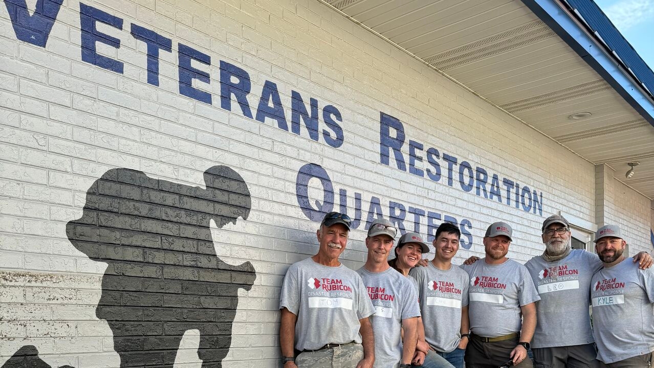 A group of volunteers wearing grey shirts that read "Team Rubicon" stand in front of a building with the words "Veterans Restoration Quarters" painted on the side.