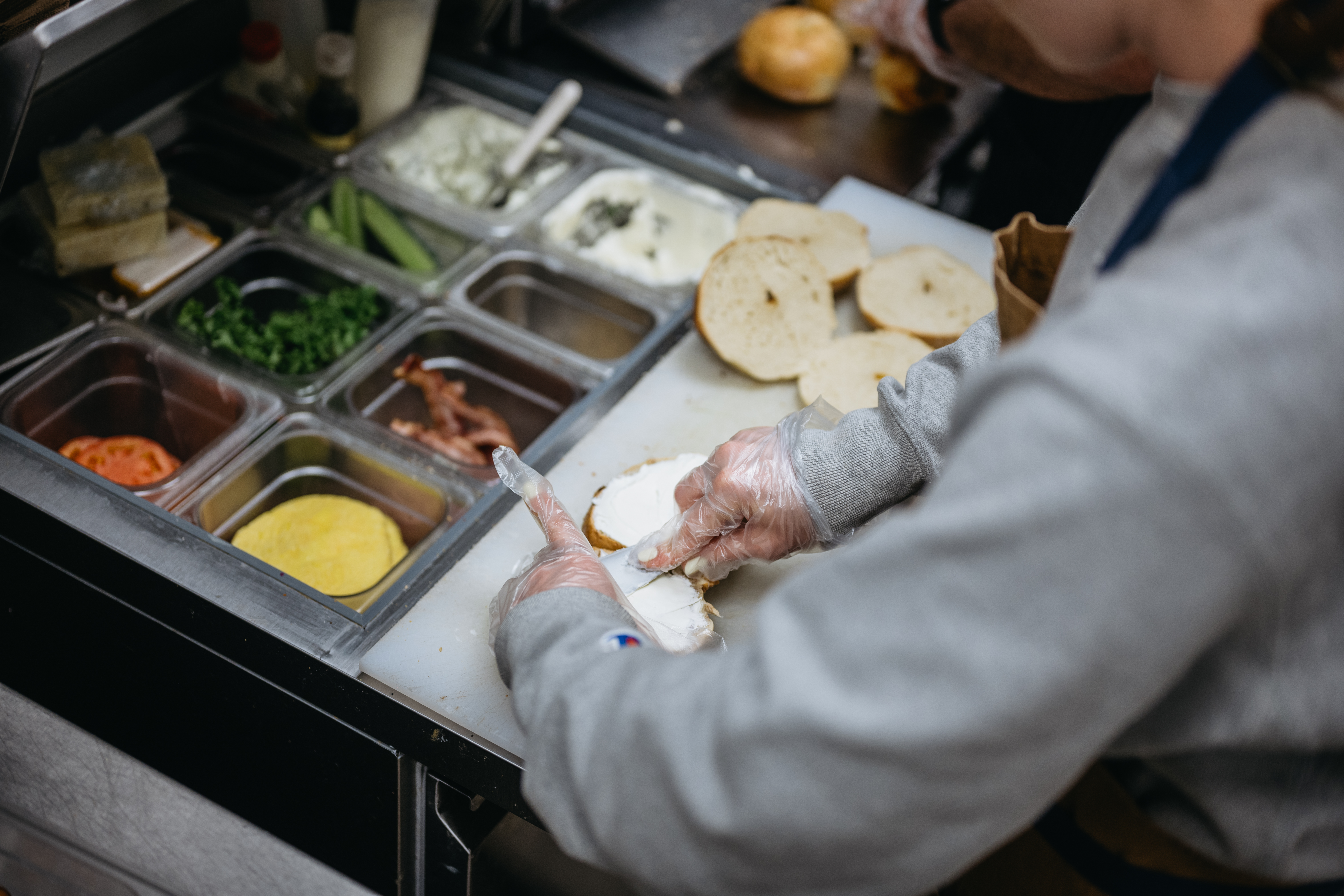 A worker at a bagel shop spreads cream cheese and toppings on a bagel.