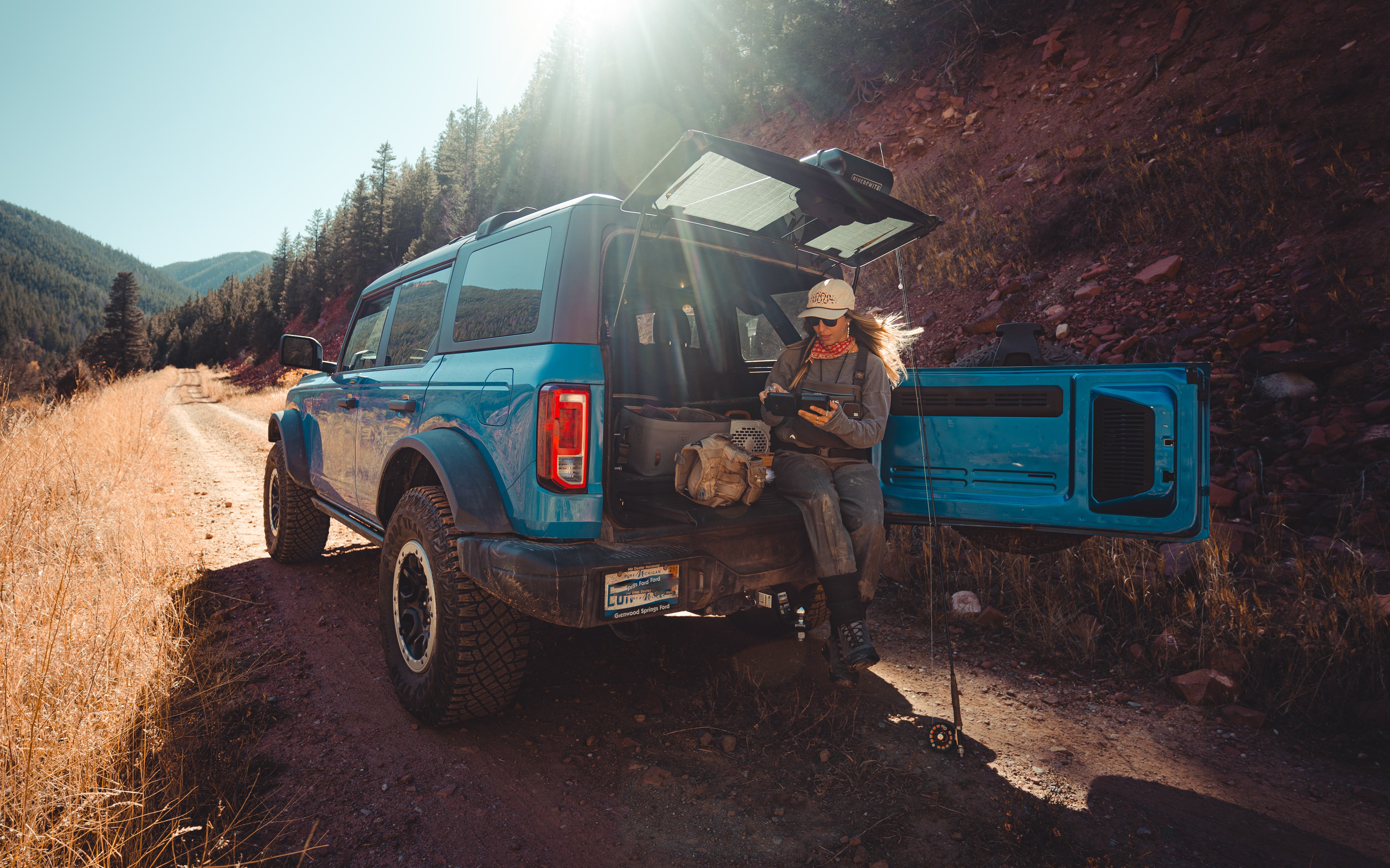 A blue Ford Bronco is parked on a dirt trail in a mountainous, forested landscape. A woman rests on the back of the Bronco with the tailgate open.
