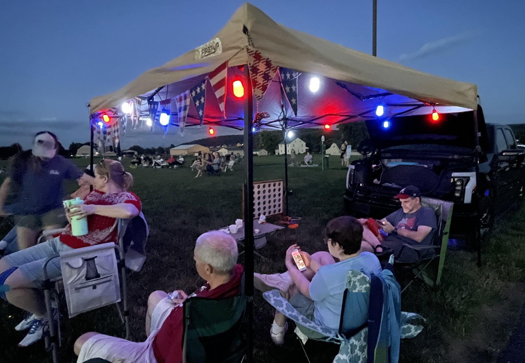 People gather around a tent decorated with red and blue lights that light up the night. Near the tent is a an electric truck with the front truck open.
