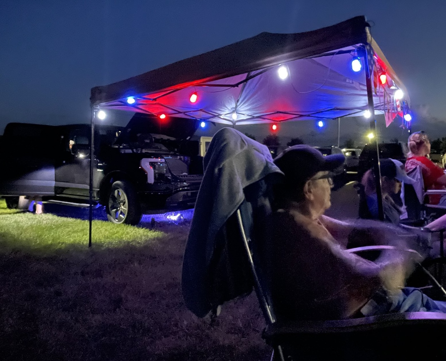 People gather around a tent decorated with red and blue lights that light up the night. Near the tent is a an electric truck with the front truck open.