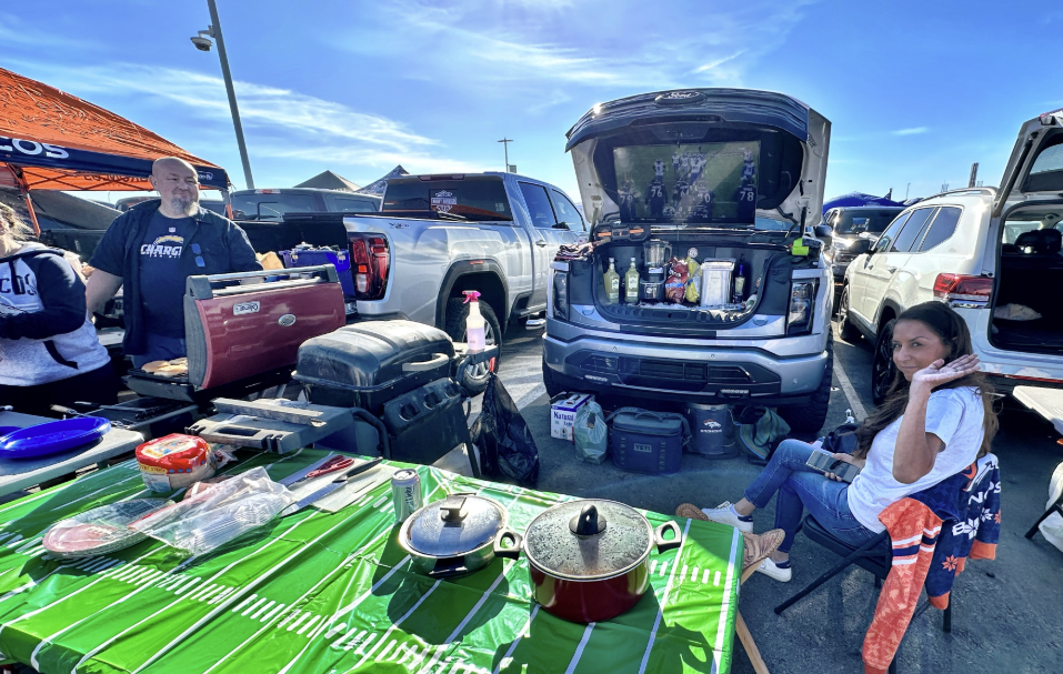 A table with food sits in front of the front trunk of an electric vehicle, which holds drinks and a tv