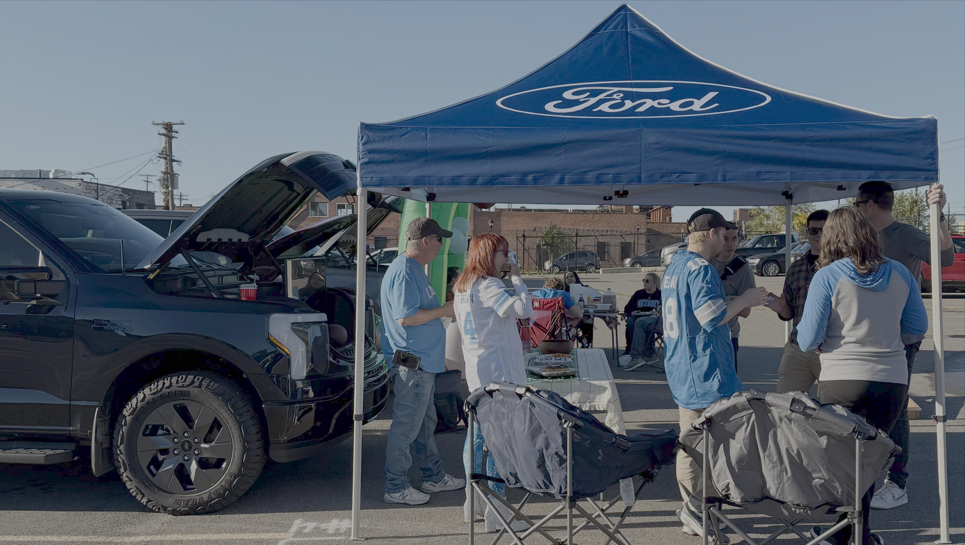 People gather under a tent with the word "Ford" on it in front of the open front trunk of an electric vehicle
