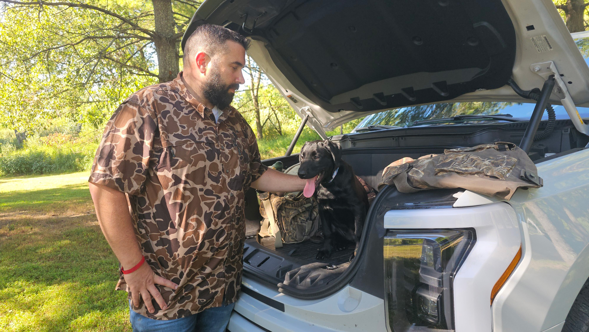 A man in a camo shirt pets his young dog, who sits in the frunk of his Ford F-150 Lightning.