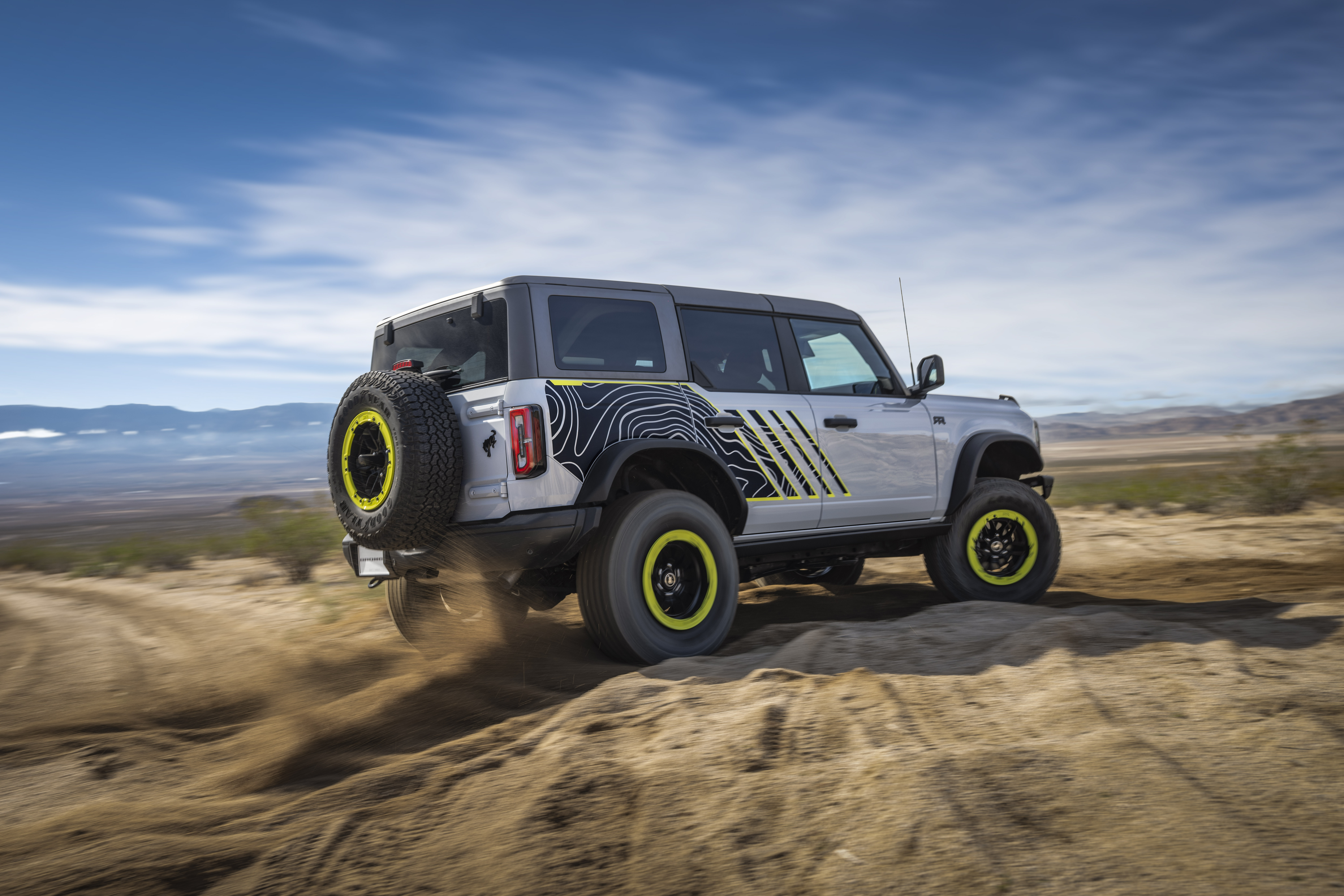 Side view of the Bronco RTR driving through sand with a blue slide and whispy clouds overhead. The vehicle is silver with lime green highlights on the wheels and side.